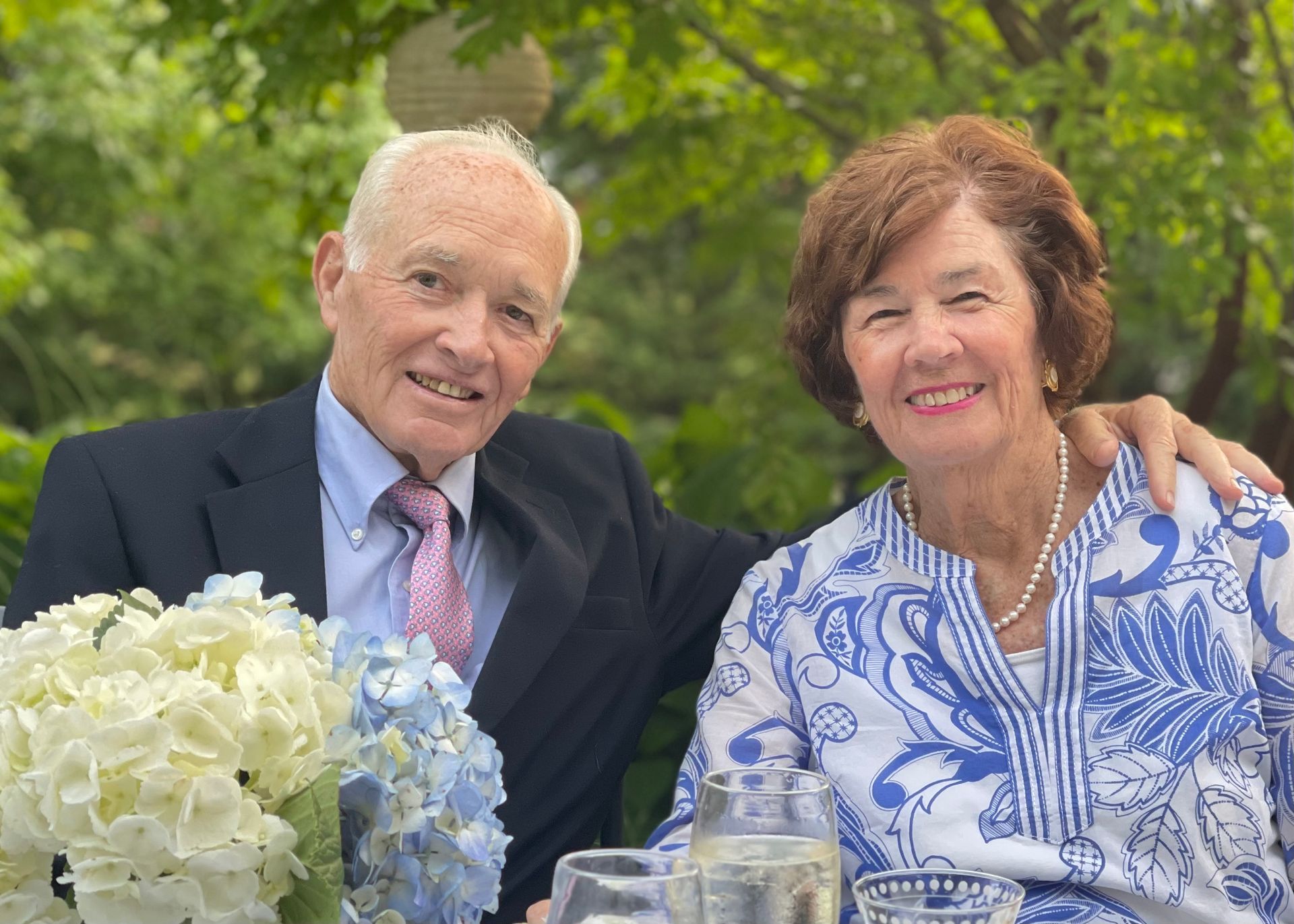 An older couple smiles together in a garden setting, with the man in a suit and the woman in a blue and white print top.