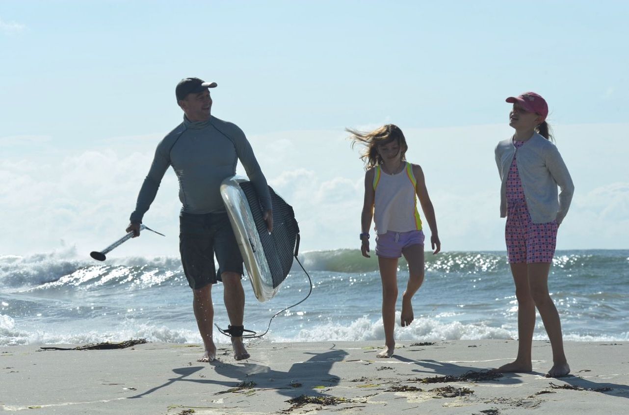 A person carrying a paddleboard walks on the beach with two children while ocean waves break in the background.