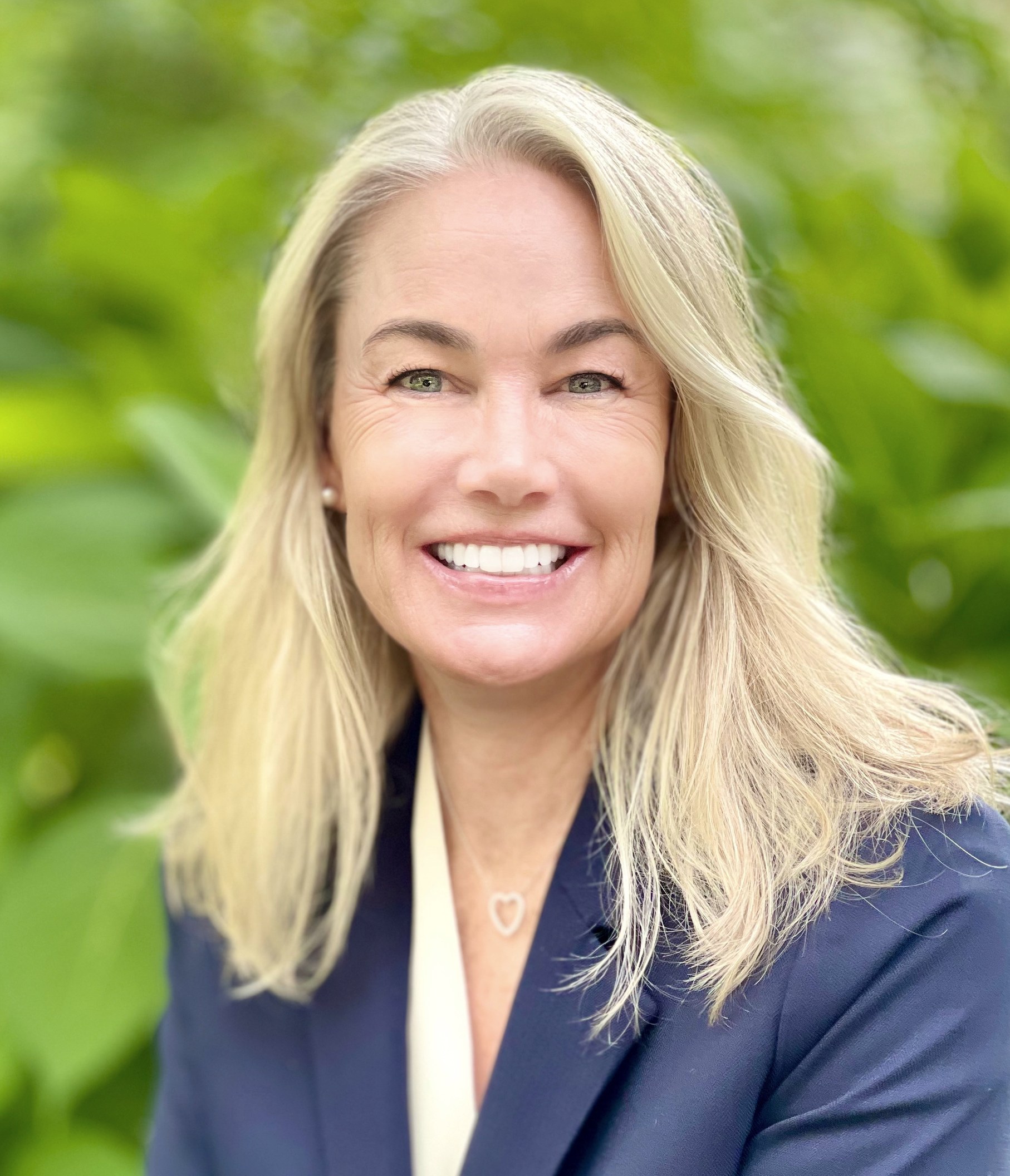 A smiling person with long blonde hair, wearing a navy blazer over a light shirt, posed against a backdrop of green leaves.