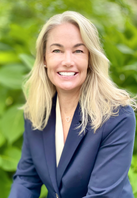 Professional portrait of a person with blonde hair smiling, wearing a blue blazer, set against a green leafy background.