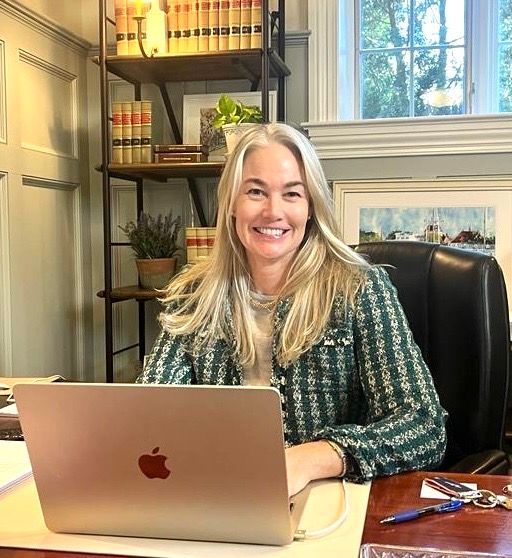 A person with long blonde hair smiles while seated at a desk with a laptop, in a room with a bookshelf and a window.