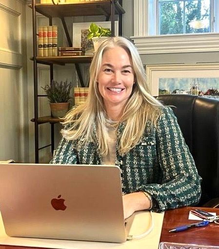 A smiling woman with long blonde hair sits at a desk using a silver laptop, with shelves of books behind her.