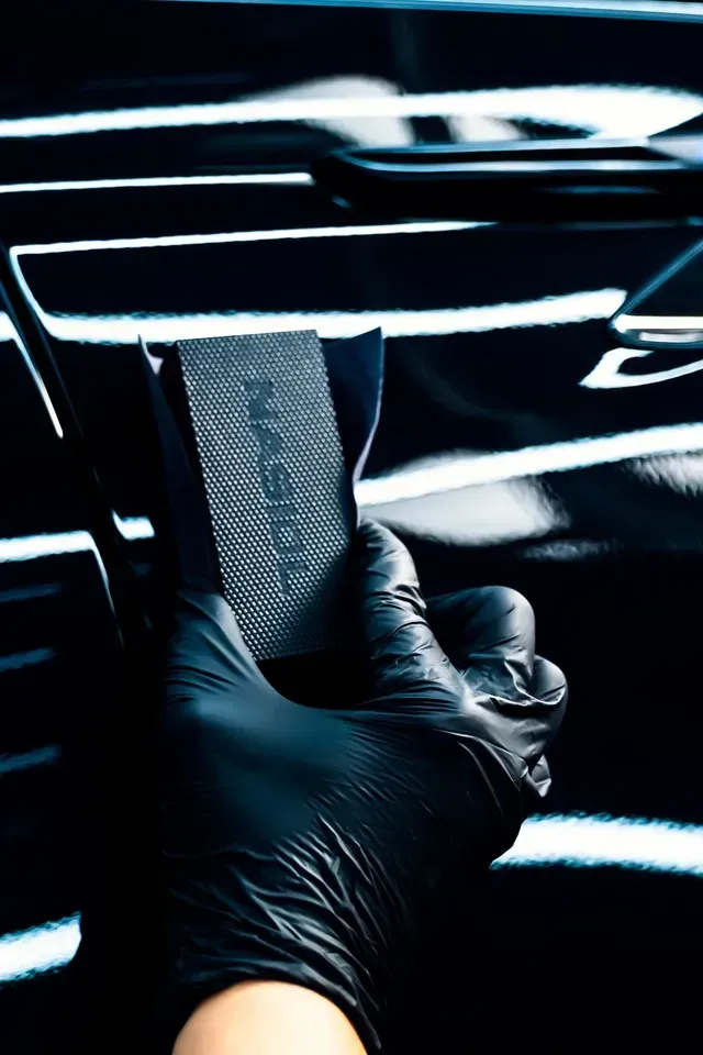 A hand in a black glove uses a foam applicator block to apply a car detailing product onto a shiny black vehicle surface.