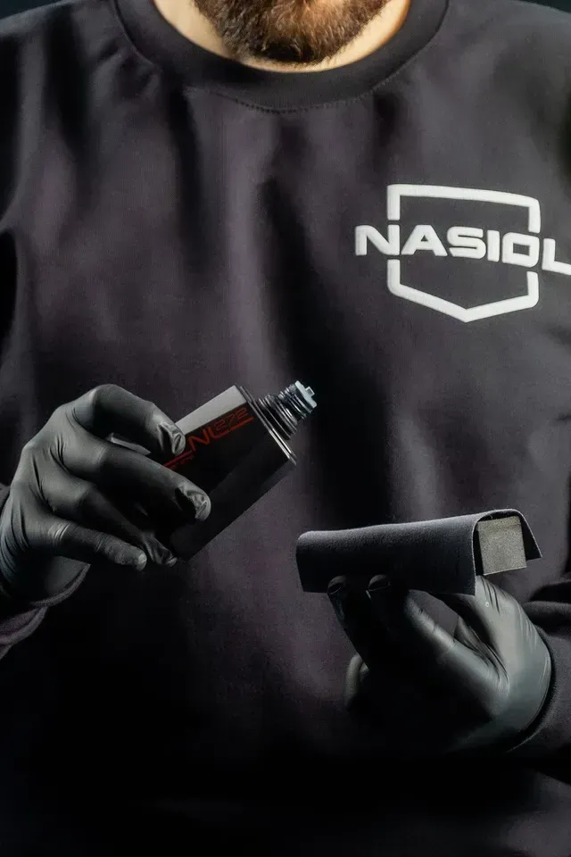 A person wearing a Nasiol shirt and black gloves applies a liquid from a bottle onto a coating applicator block.