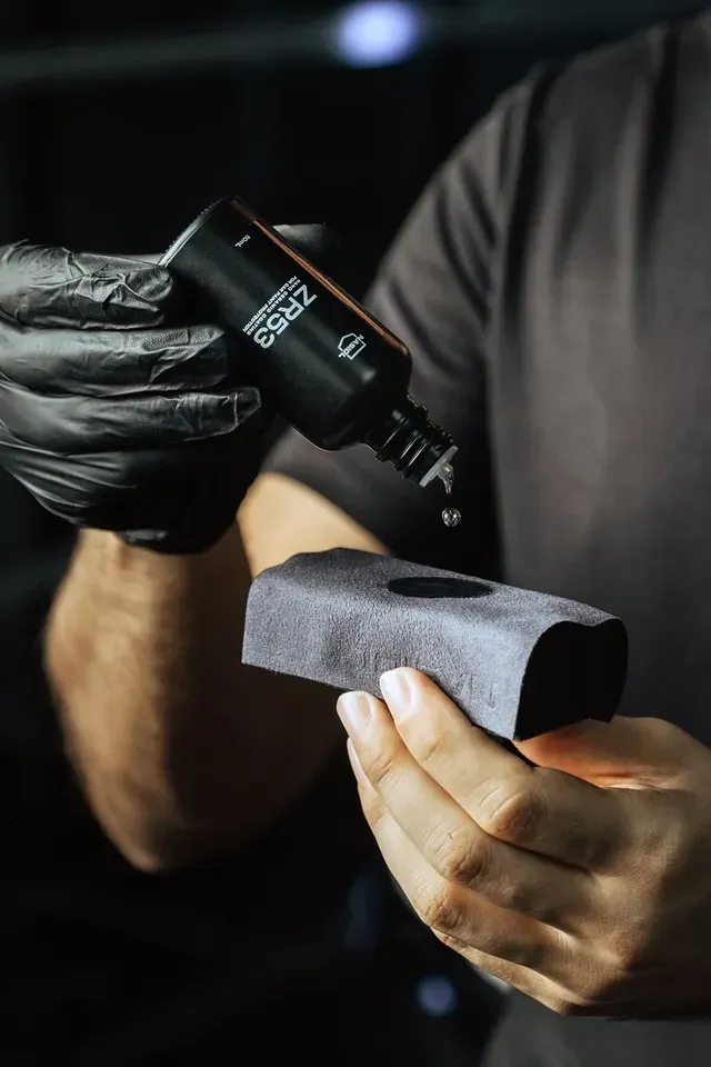 A person wearing a black glove applying liquid from a small black bottle onto a grey applicator pad.