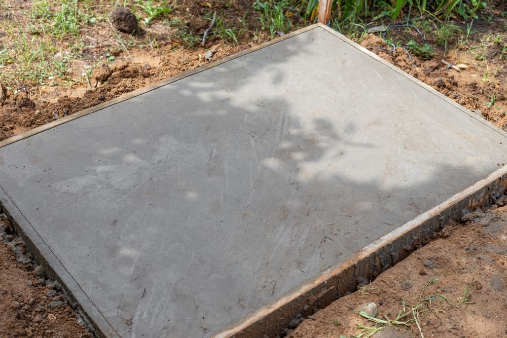 Freshly poured rectangular concrete slab in a dirt yard, with shadows from nearby trees.