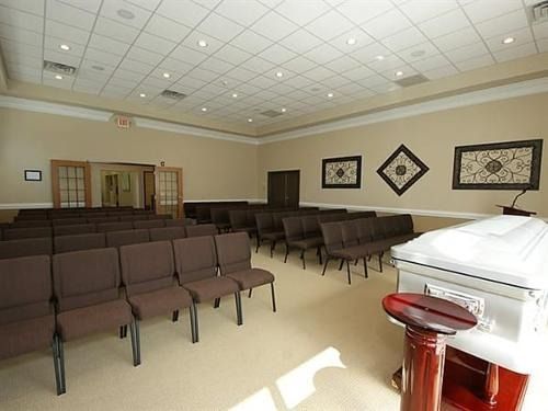 Empty waiting room with rows of brown chairs, beige walls, framed art, and a white casket at the front.