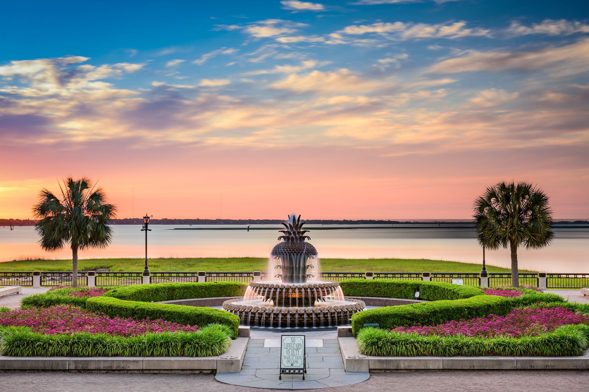 Sunset garden with a central fountain, flower beds, palm trees, and a calm waterfront beyond.