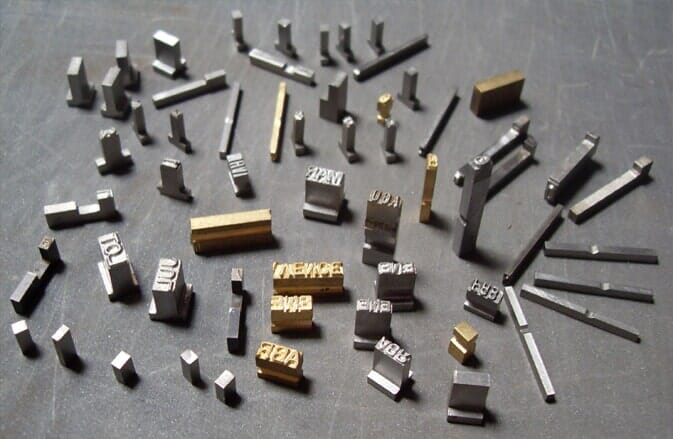 Piezas de tipos de metal esparcidas sobre una superficie gris, incluyendo letras, números y formas para imprimir.