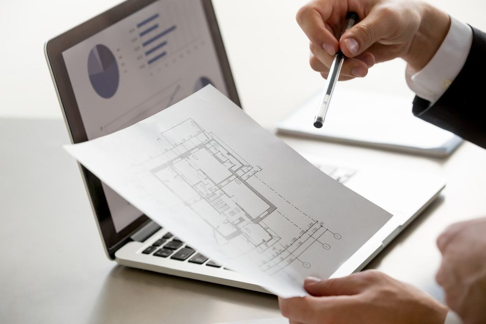 Person pointing at blueprint, next to laptop displaying graphs.