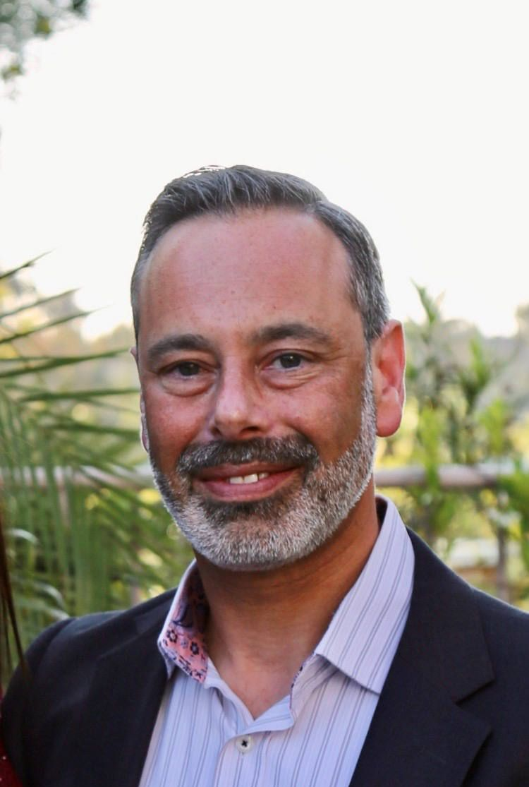 Smiling man in a dark blazer and striped shirt outdoors with greenery behind him