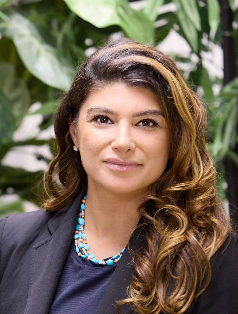 Portrait of a woman in a dark blazer and turquoise necklace, standing before leafy greenery