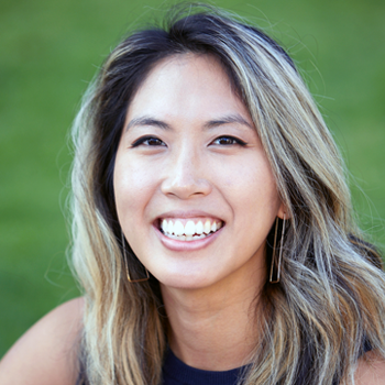 Smiling person with long highlighted hair outdoors against a blurred green background