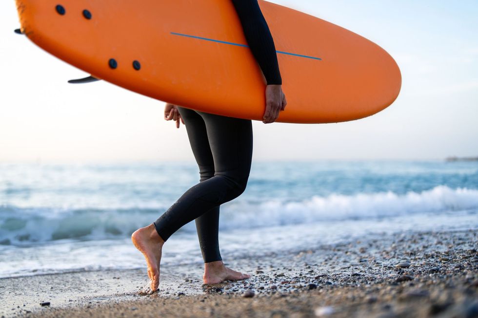 Surfer carrying an orange surfboard on a pebbled beach beside the ocean