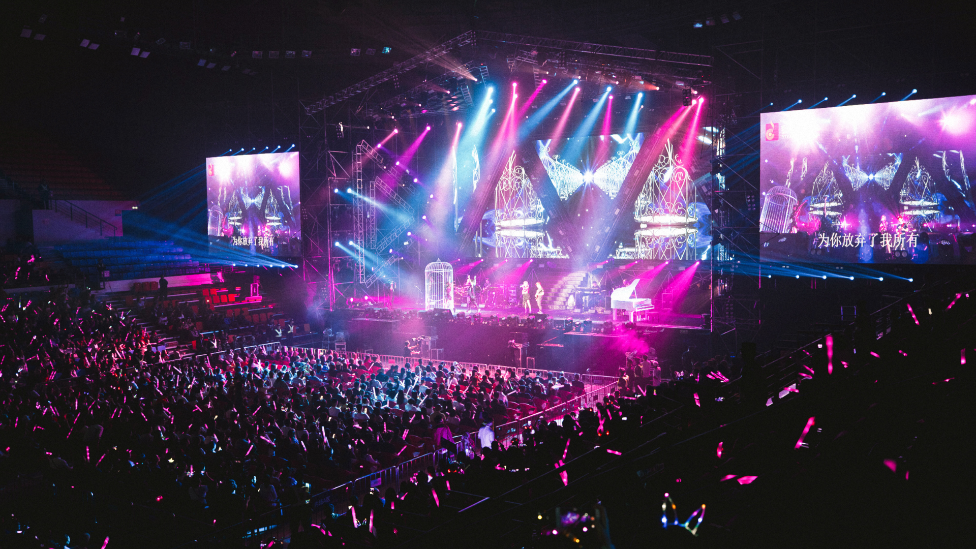 Concert stage with bright blue and pink lights, cheering crowd with glowing wands.