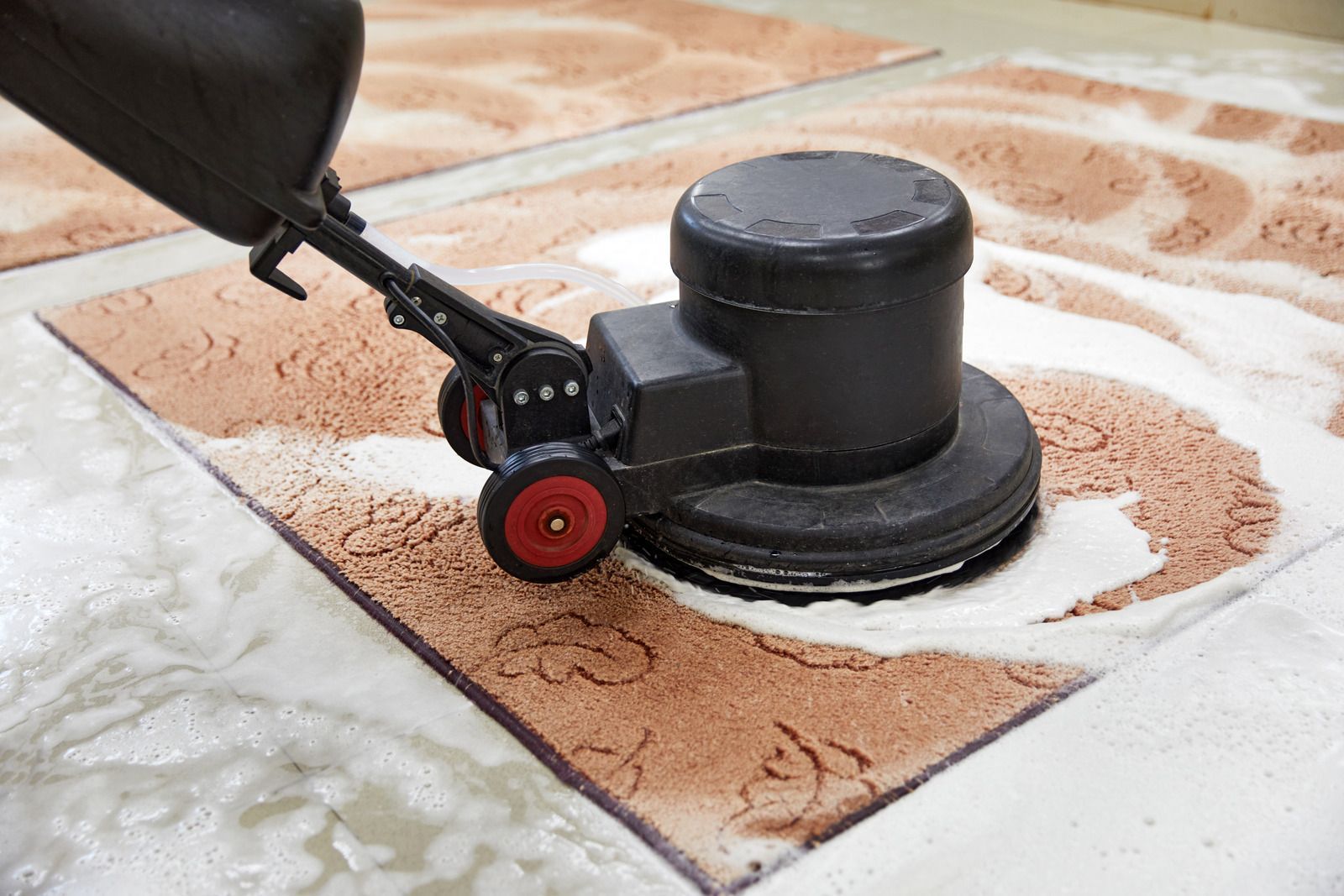 A black floor buffer cleans a brown, patterned rug covered in white foam.