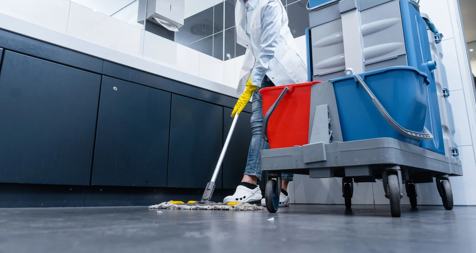 Janitor mopping a clean floor beside a blue cleaning cart in a modern hallway