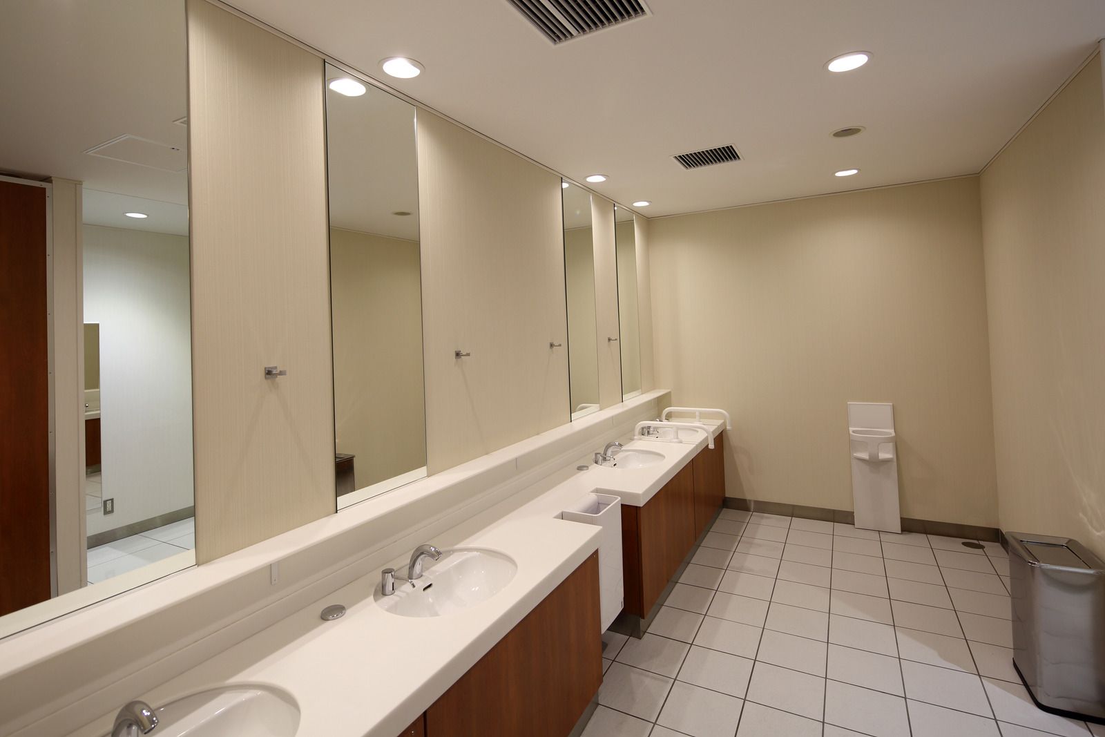 Public restroom with beige walls, long mirrors, and a row of white sinks.