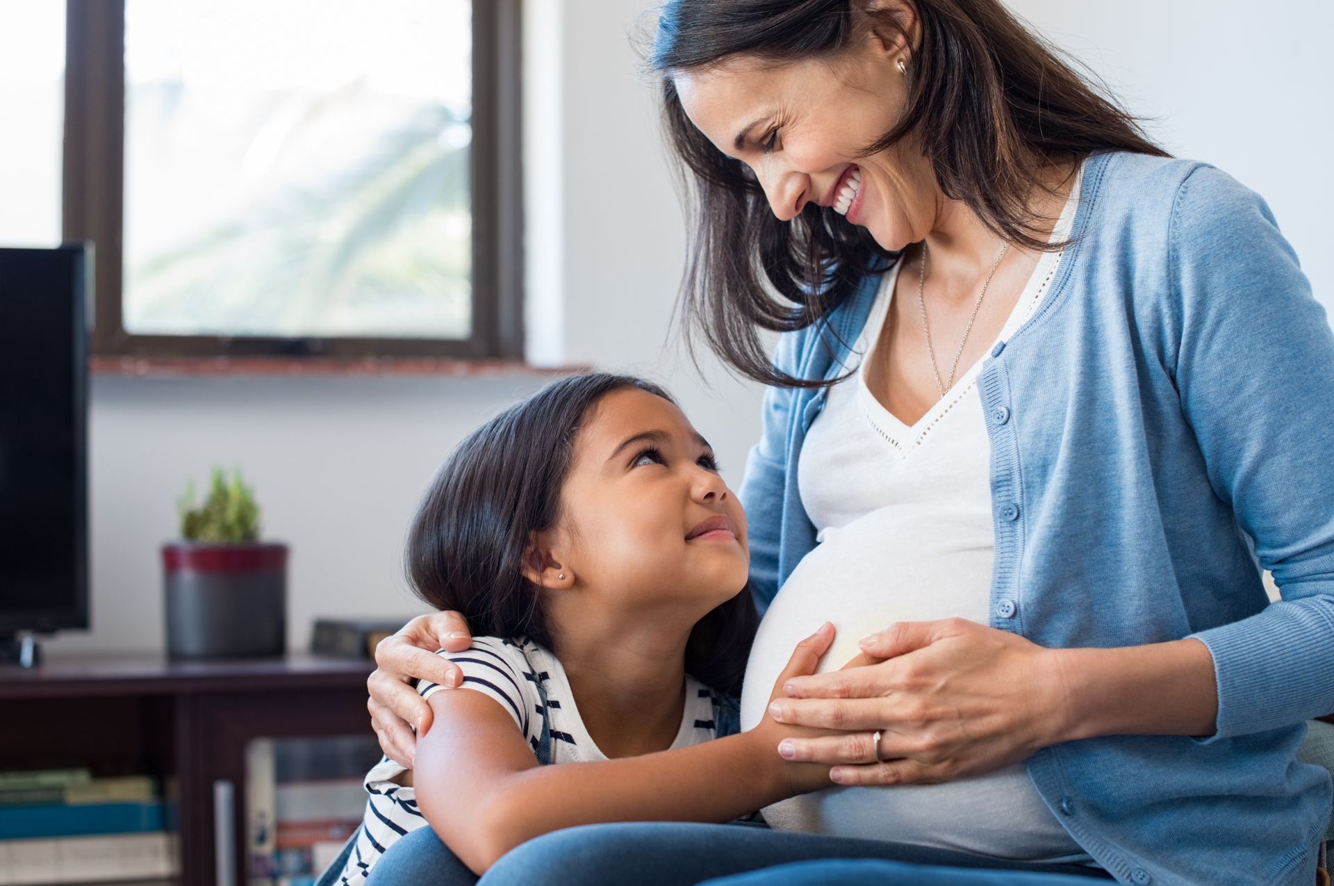 Pregnant woman smiling at a child touching her belly indoors.