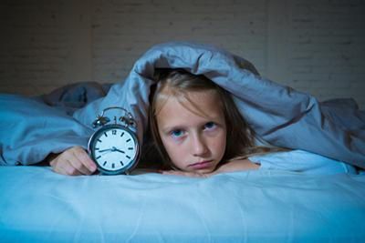 A girl is lying under blankets with an alarm clock in her hand