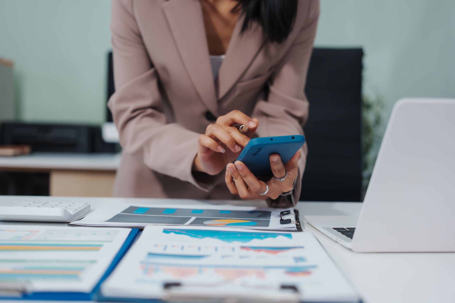 Woman in a blazer using a phone, working at a desk with charts and a laptop. Woman in a blazer using a phone, working at a desk with charts and a laptop.