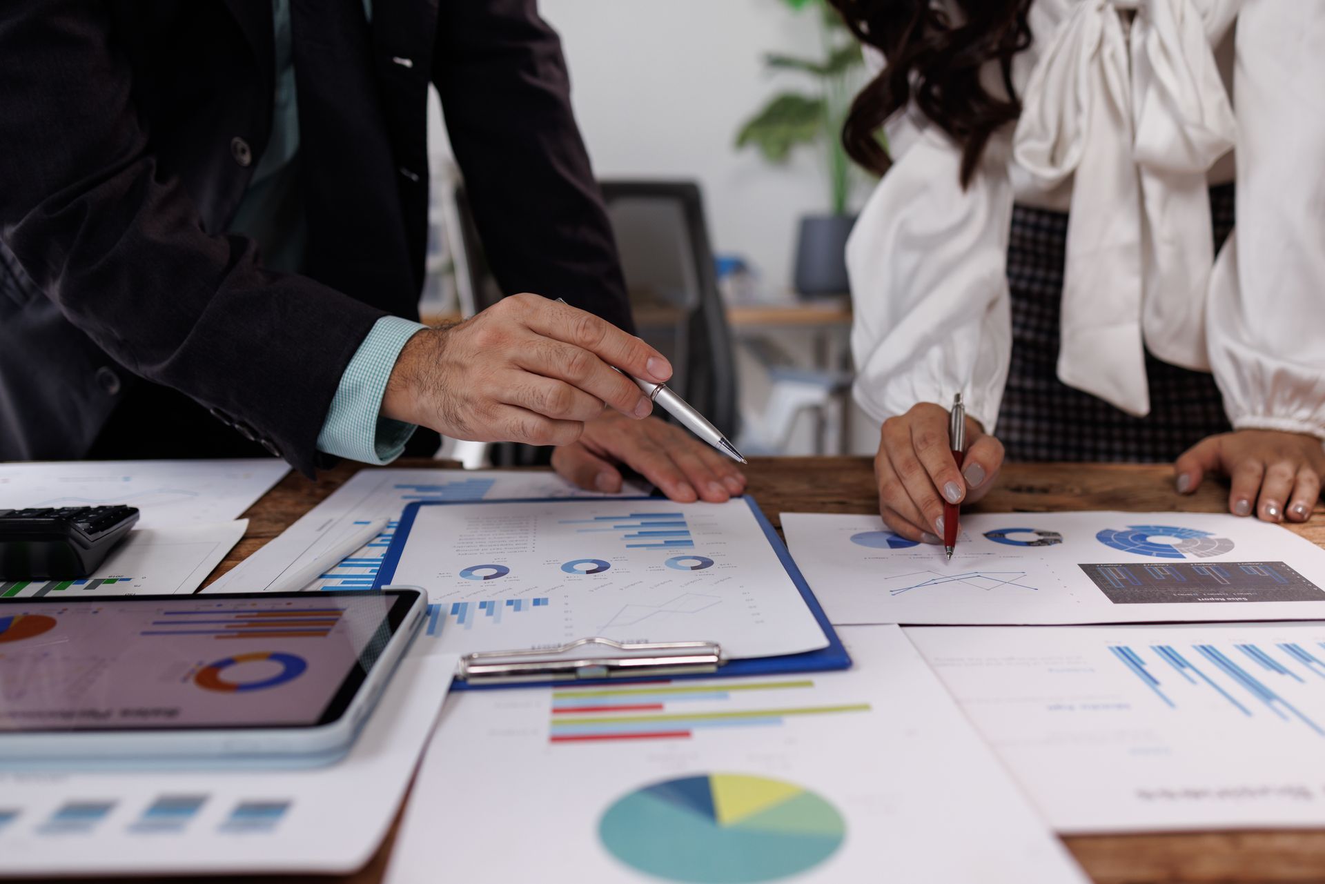 Two people reviewing financial documents, pointing at charts and graphs on a wooden table. Two people reviewing financial documents, pointing at charts and graphs on a wooden table.