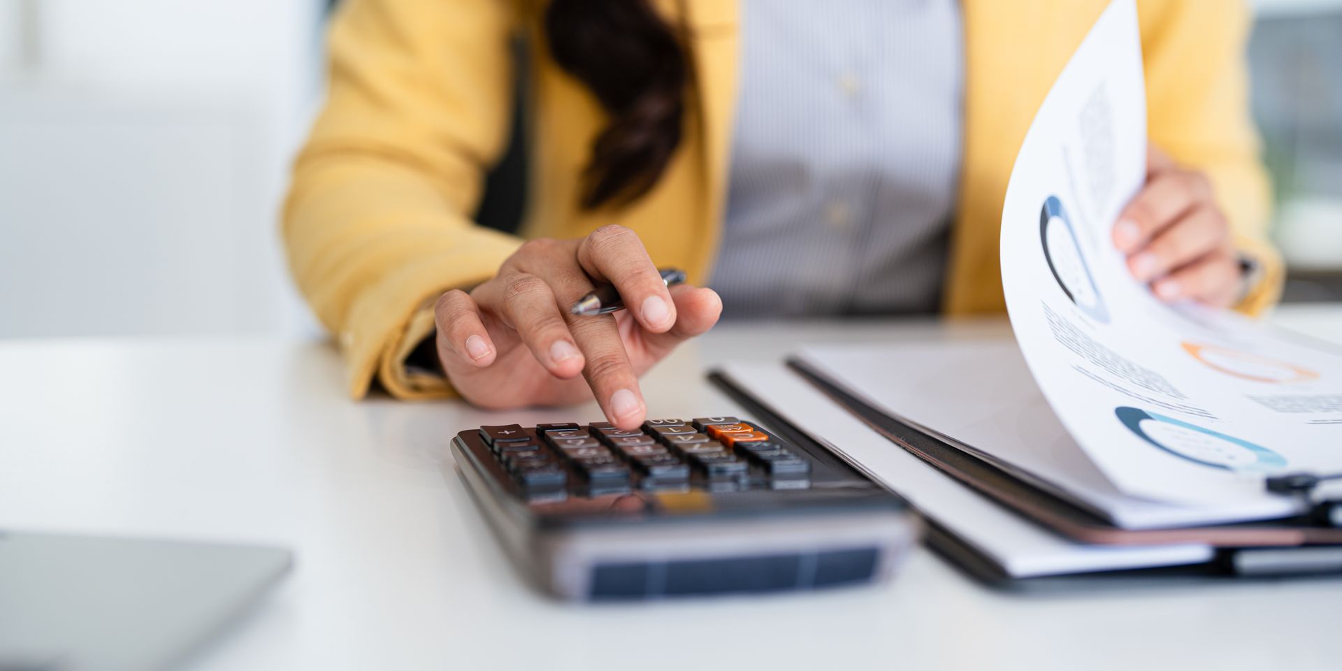 Person in yellow blazer using calculator and reviewing papers with charts.