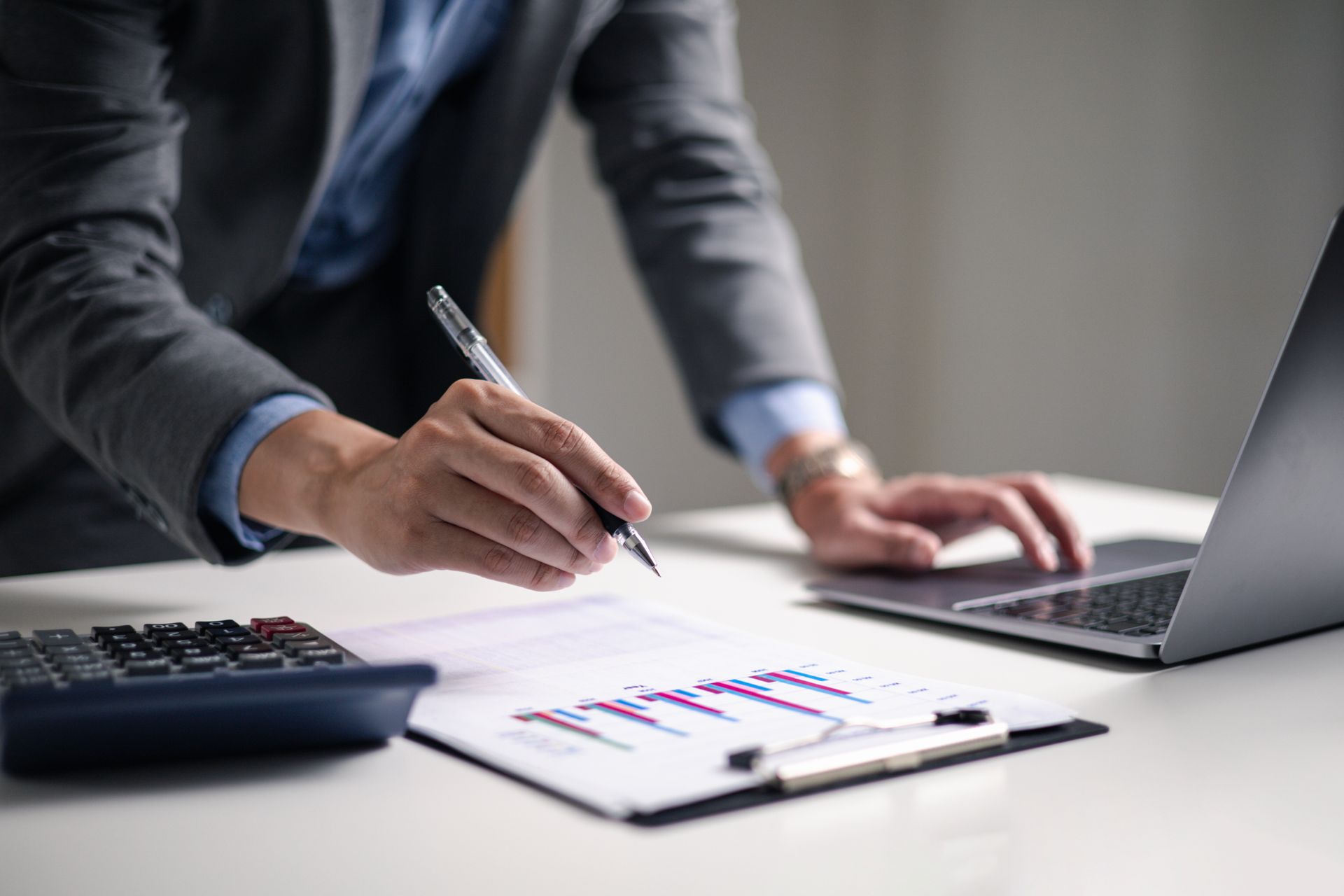 Person in a suit writing on a chart, using a laptop and calculator at a white desk.