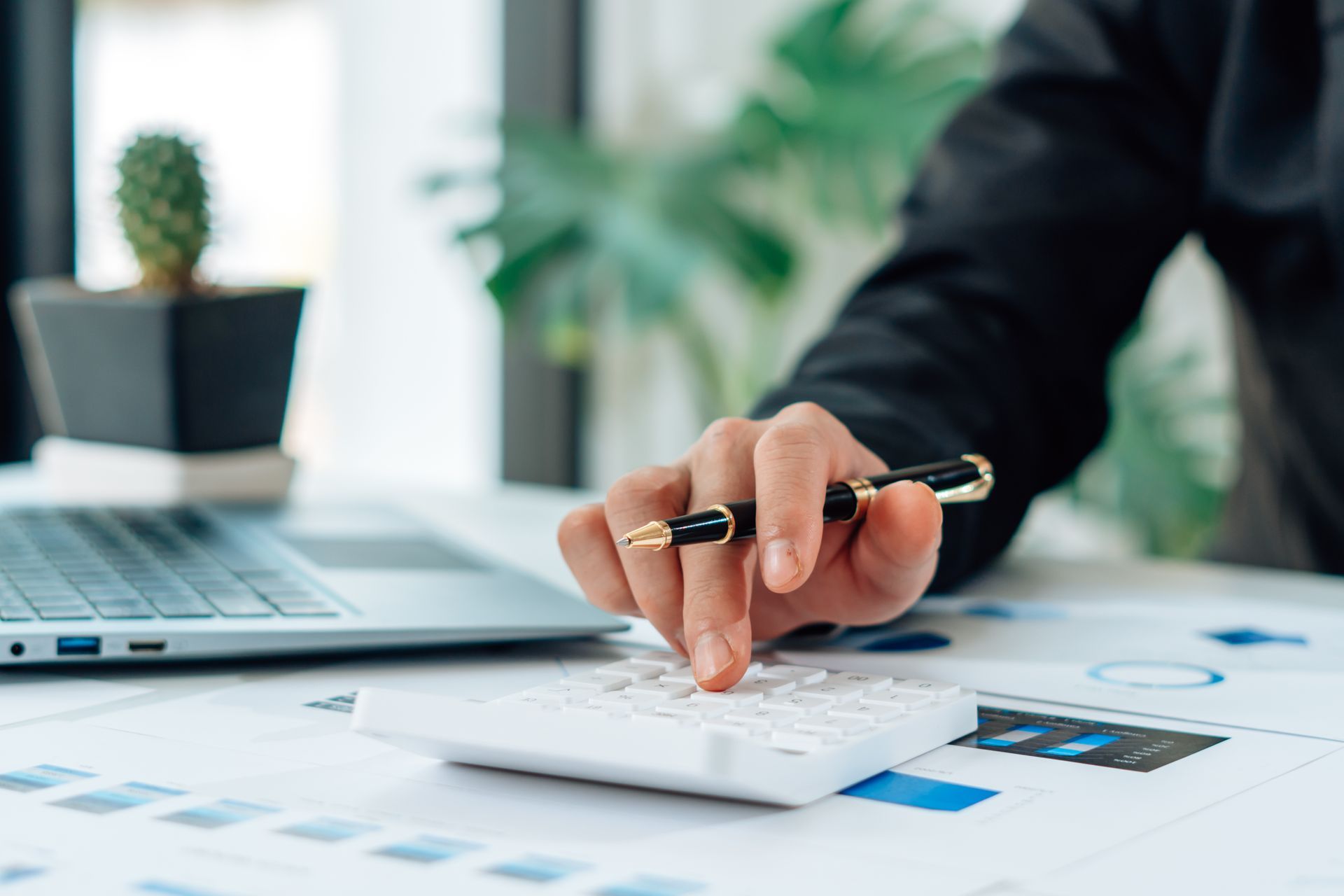 Person in black suit using calculator at desk with laptop, financial documents, and cactus.