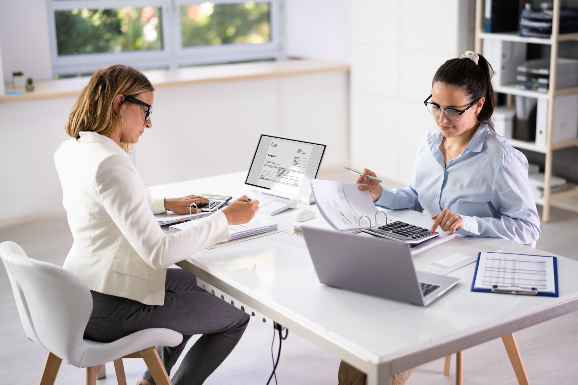 Two women working at a white desk with calculators, paperwork, and laptops in an office. Two women working at a white desk with calculators, paperwork, and laptops in an office.