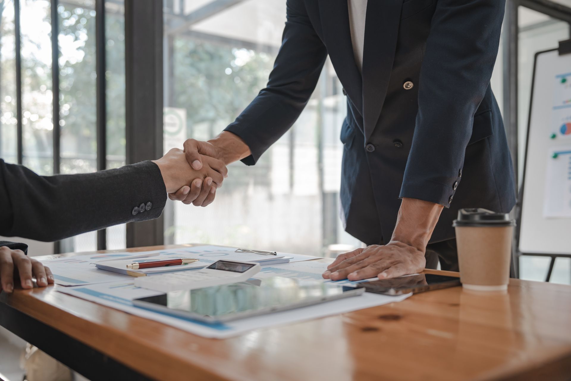 Two people in suits shaking hands over a table with papers, a tablet, and a coffee cup. Two people in suits shaking hands over a table with papers, a tablet, and a coffee cup.