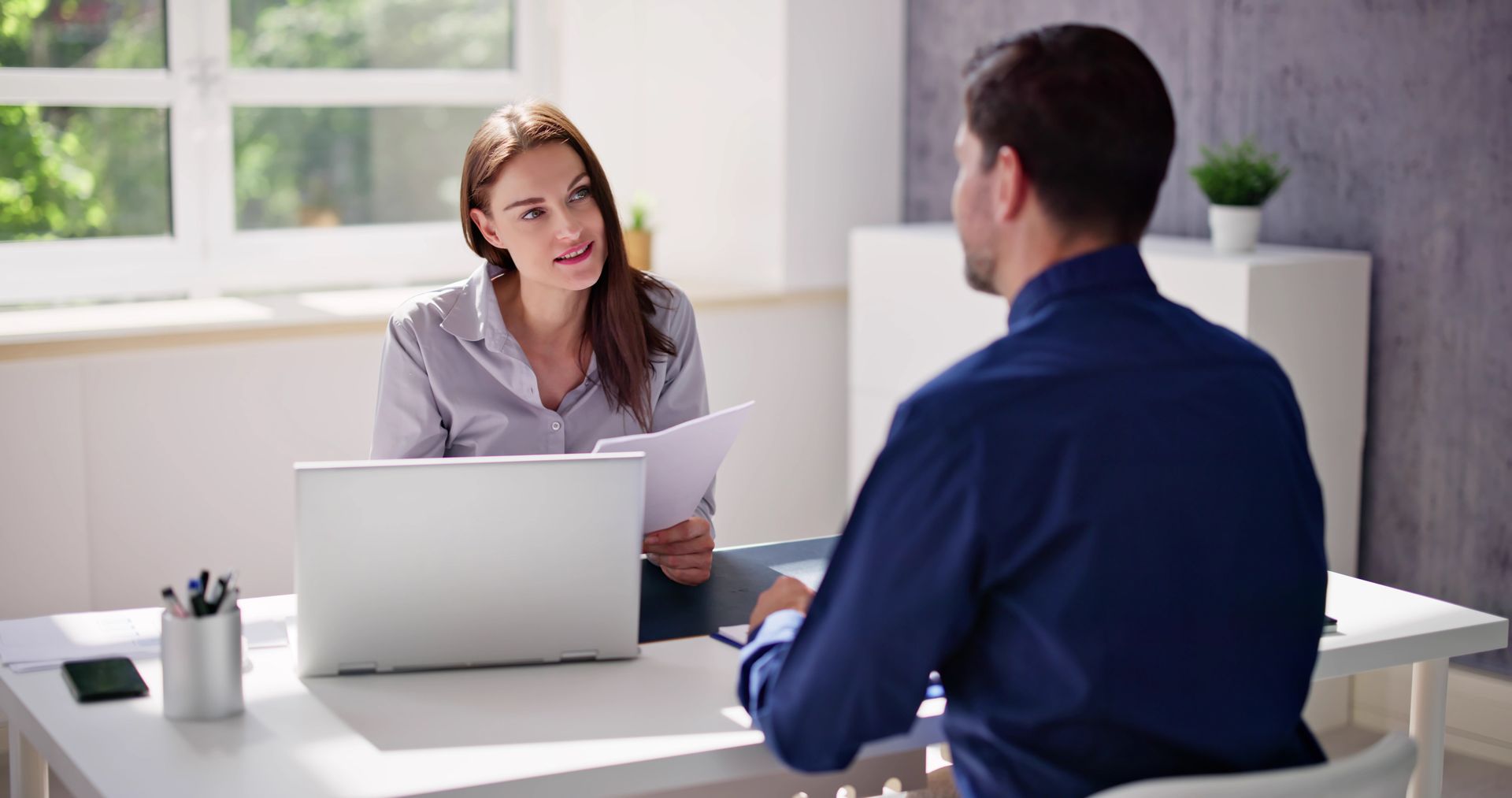 Woman interviewing a person seated at a table, holding papers, with a laptop.