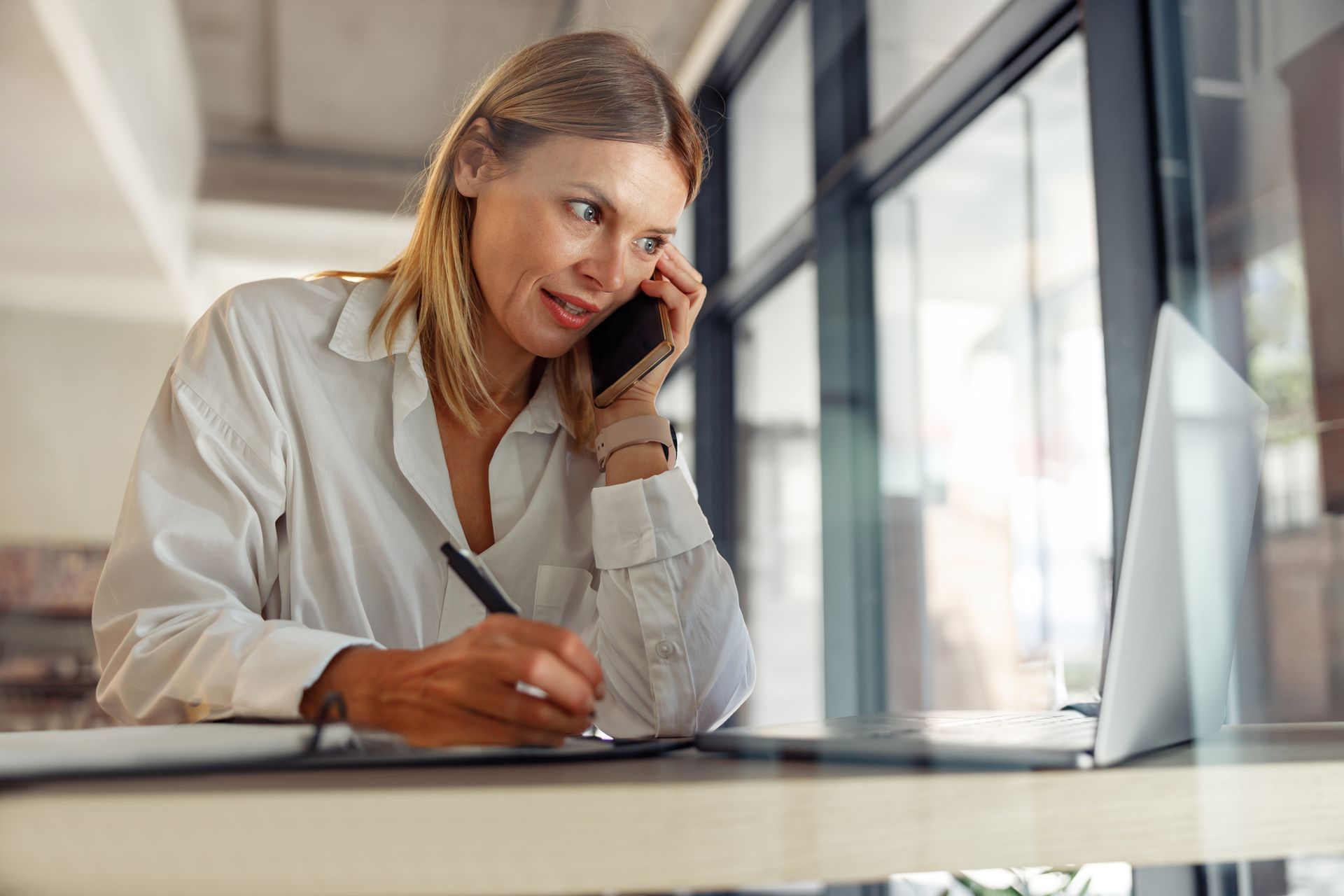 Woman on phone, writing notes, working at desk with laptop in front of a window. Woman on phone, writing notes, working at desk with laptop in front of a window.