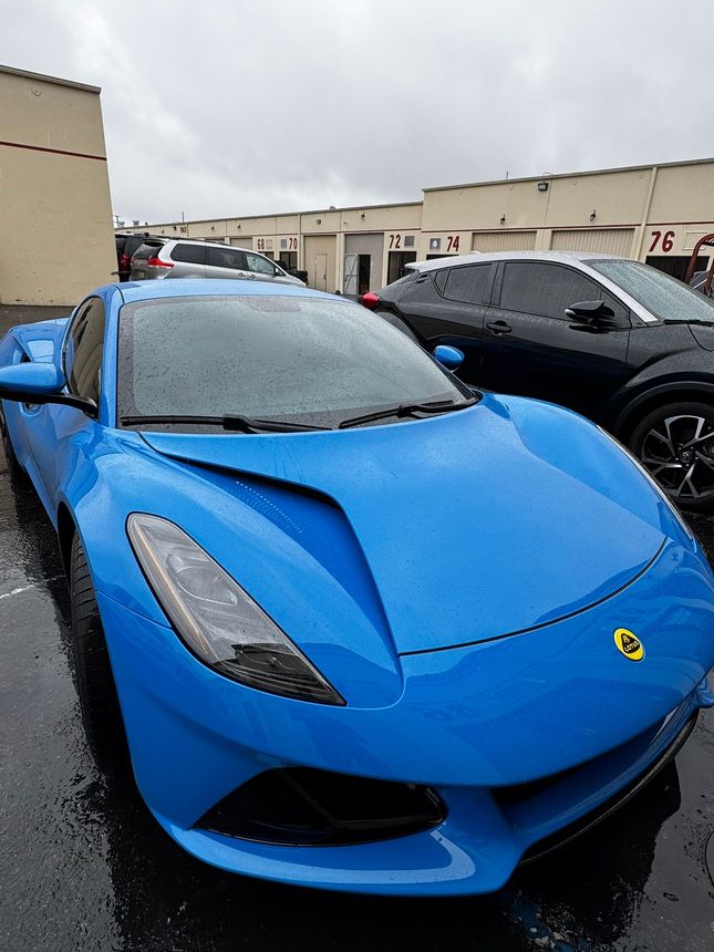 Bright blue sports car parked outside, rain-covered asphalt. Black car in the background.