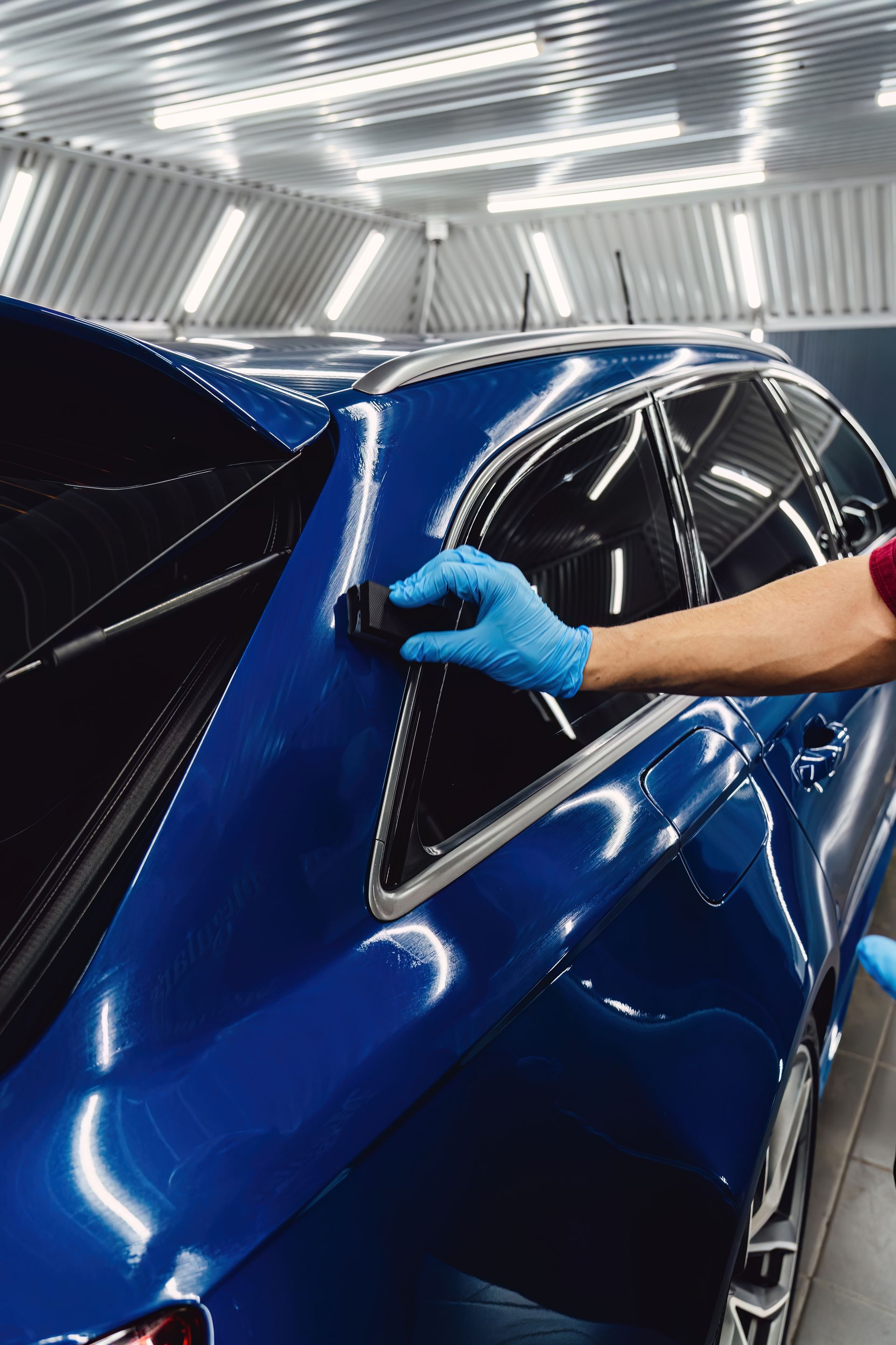 Person wearing blue gloves applying something to a blue car in a well-lit shop.