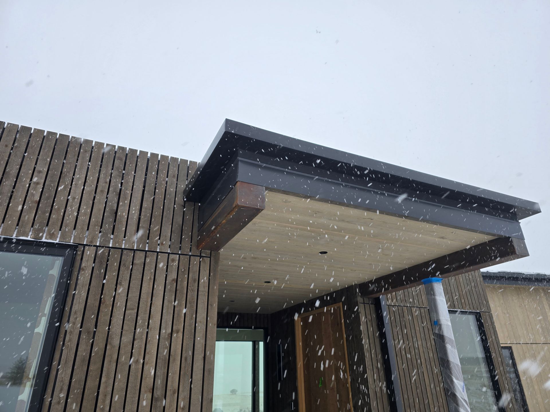 A contemporary building exterior with vertical wood slat siding and a dark roof overhang during a light snowfall.