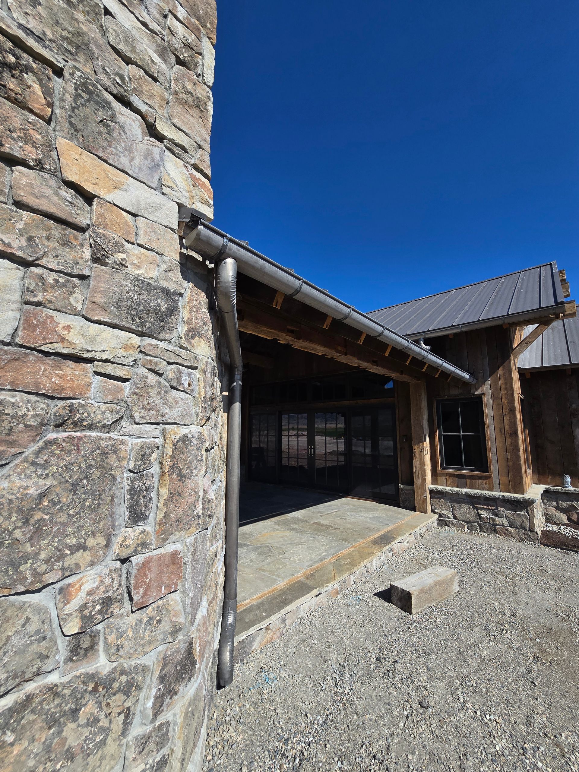 A stone wall and roof overhang of a rustic building with a metal gutter, set against a bright blue sky.