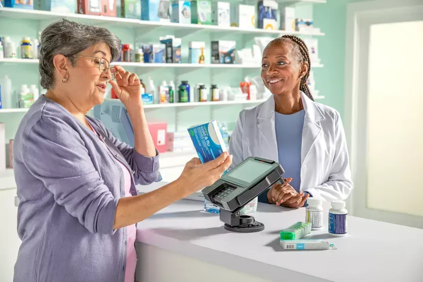 Two people at a pharmacy counter, one holding a medicine box while speaking with a smiling pharmacist.