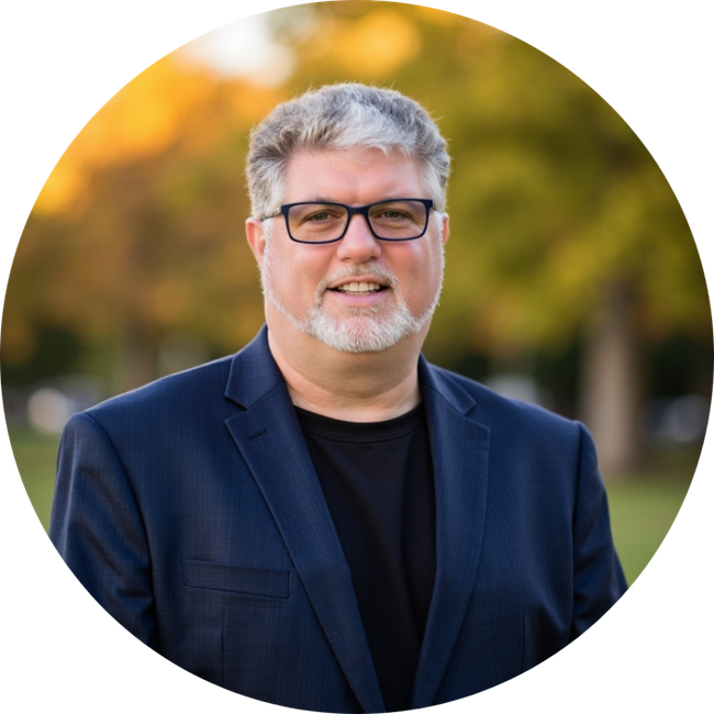 Smiling man in glasses and navy blazer standing outdoors with blurred autumn trees behind him