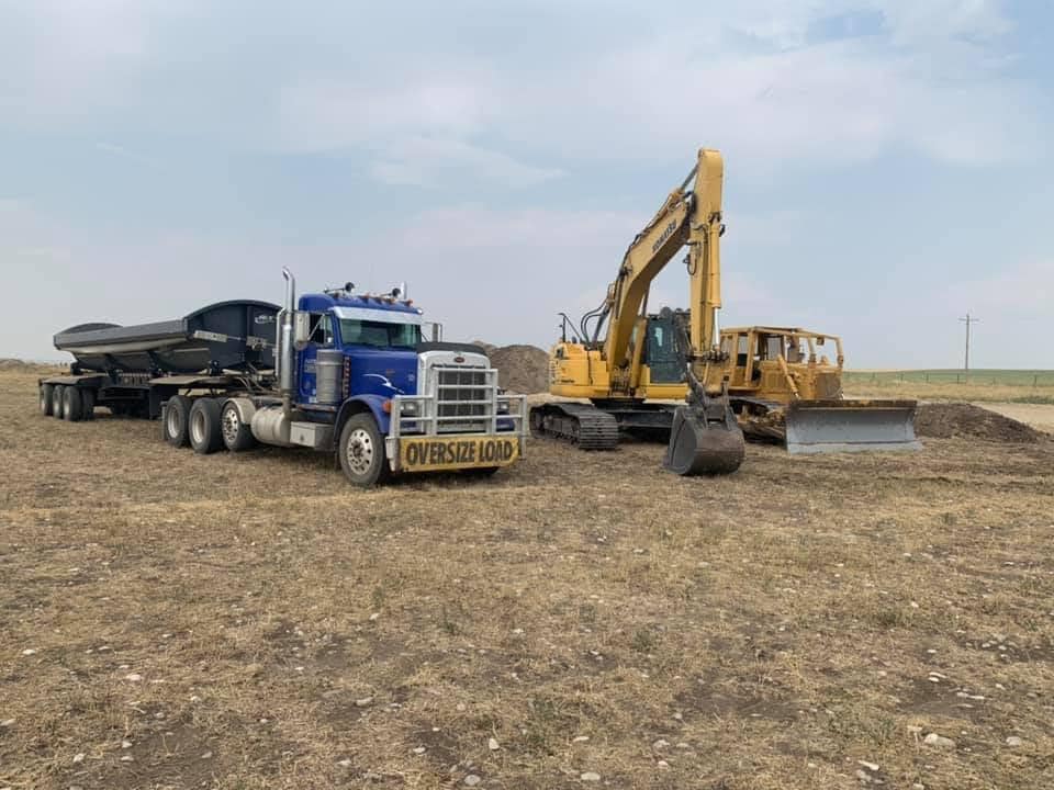 Blue semi-truck with trailer and yellow excavator on dirt field under cloudy sky.