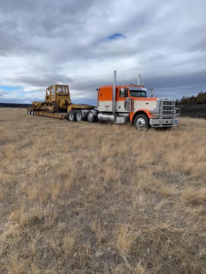 Orange and white semi-truck pulling a yellow bulldozer on a flatbed trailer across a grassy field under a cloudy sky.