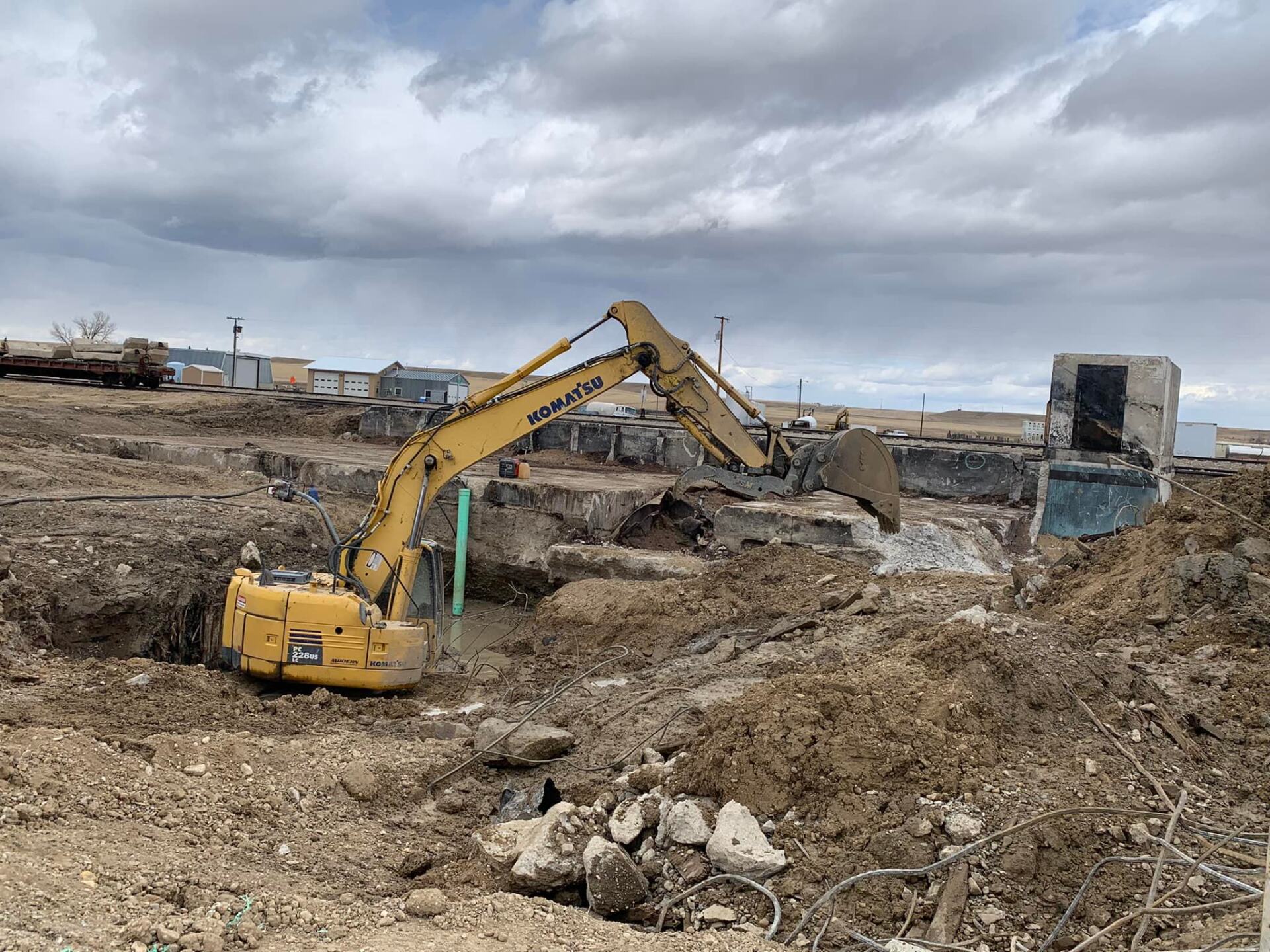 Yellow excavator demolishing concrete structure in a construction site, under cloudy sky.