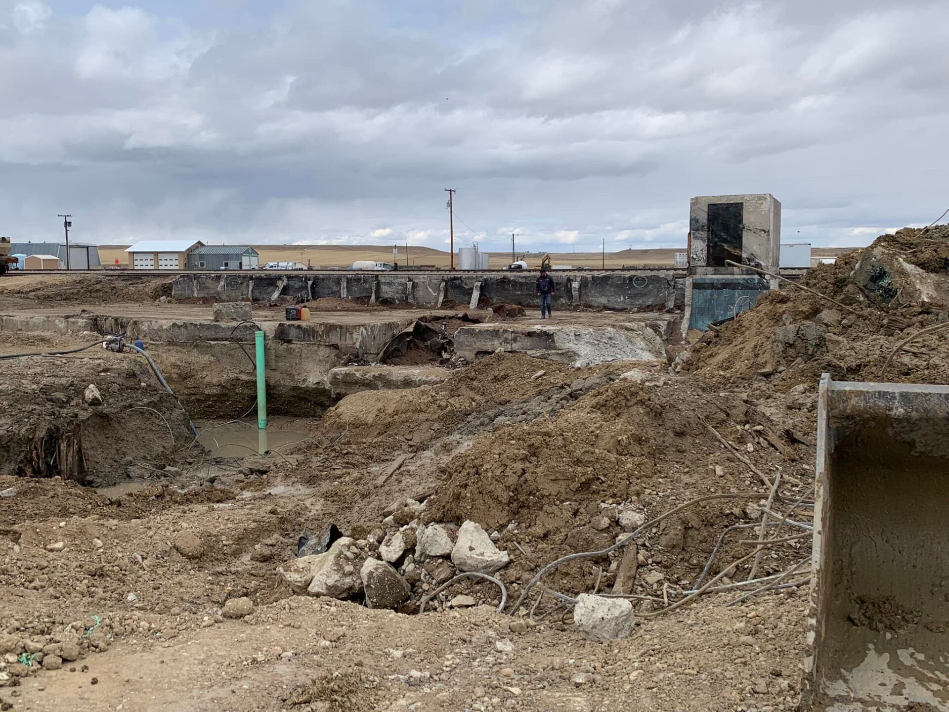 Construction site with excavated earth, concrete structures, and workers under a cloudy sky.