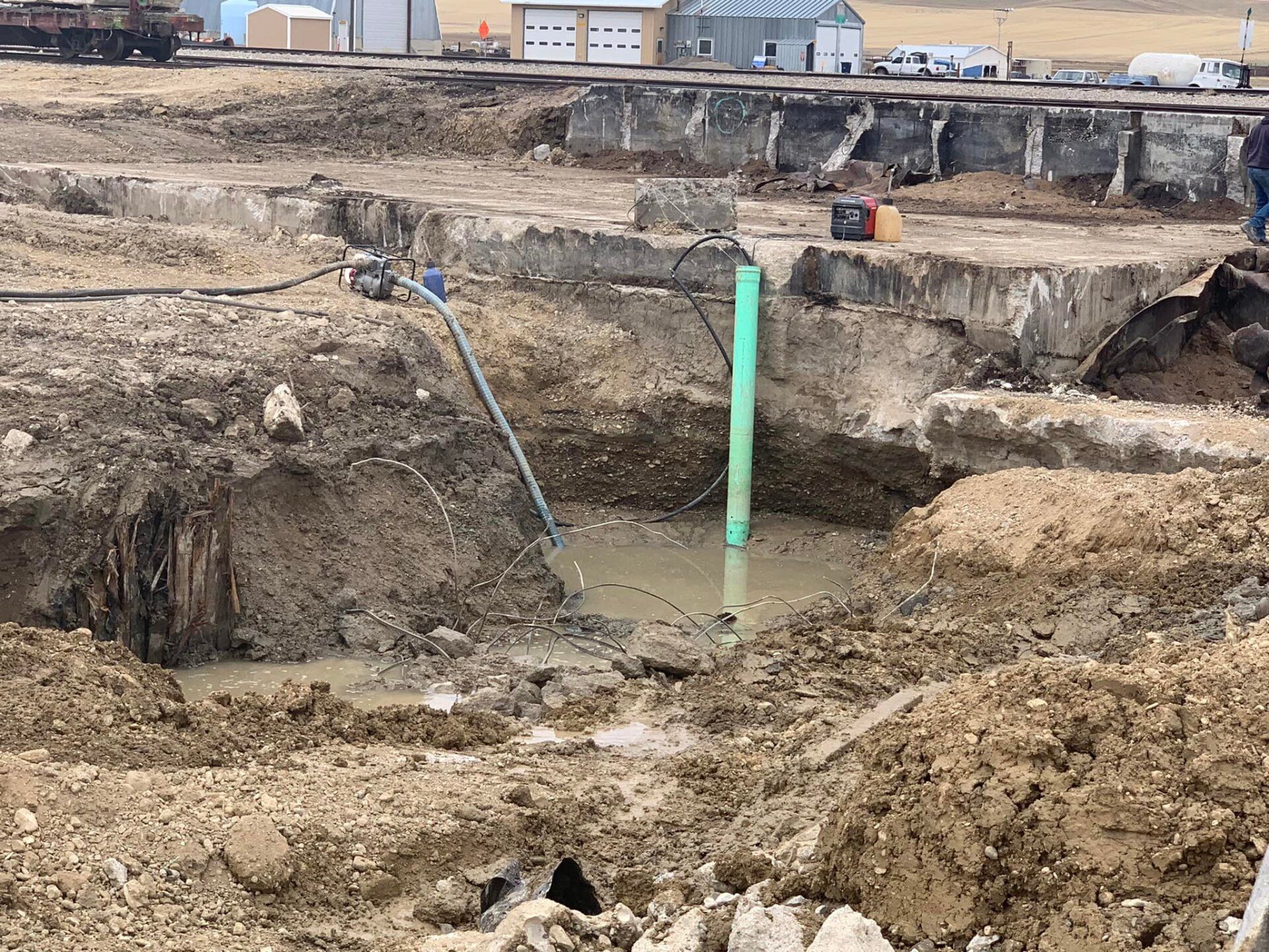 Construction site with excavation pit and standing water. A green pipe stands in the muddy pit.