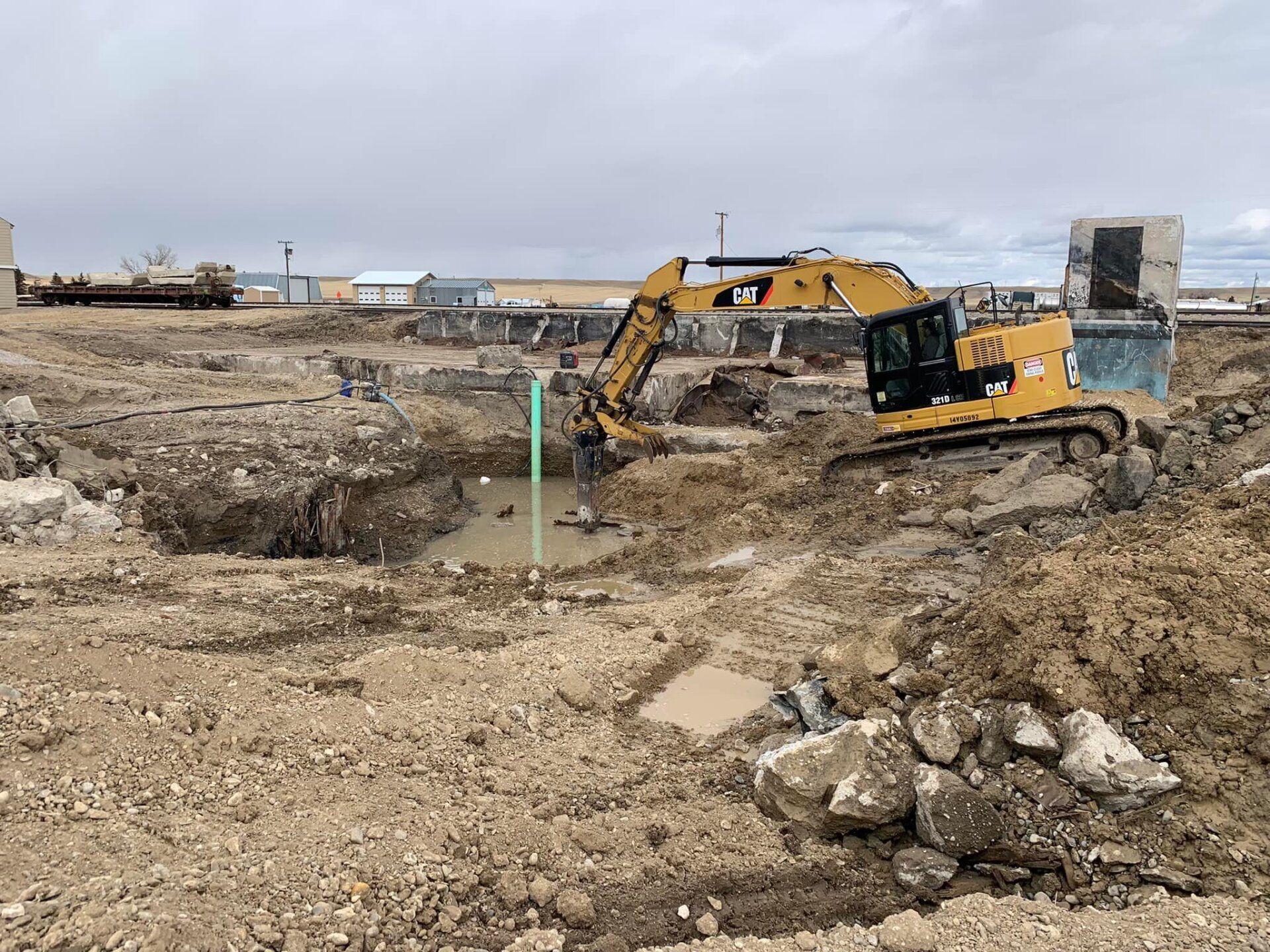 An excavator digs in a construction site with muddy ground and some existing structures.