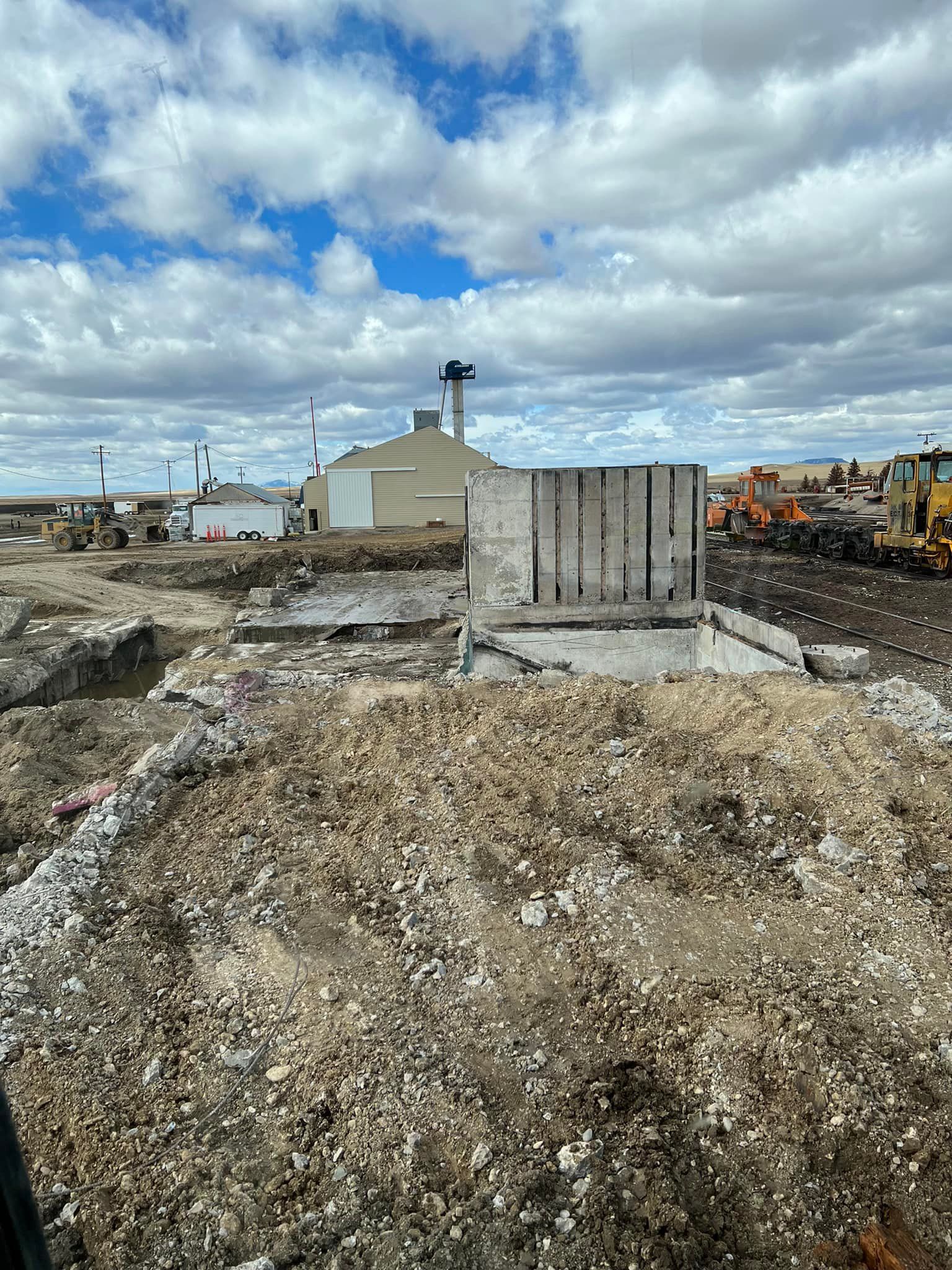 Construction site with partially demolished concrete structure; beige building and heavy machinery in background.