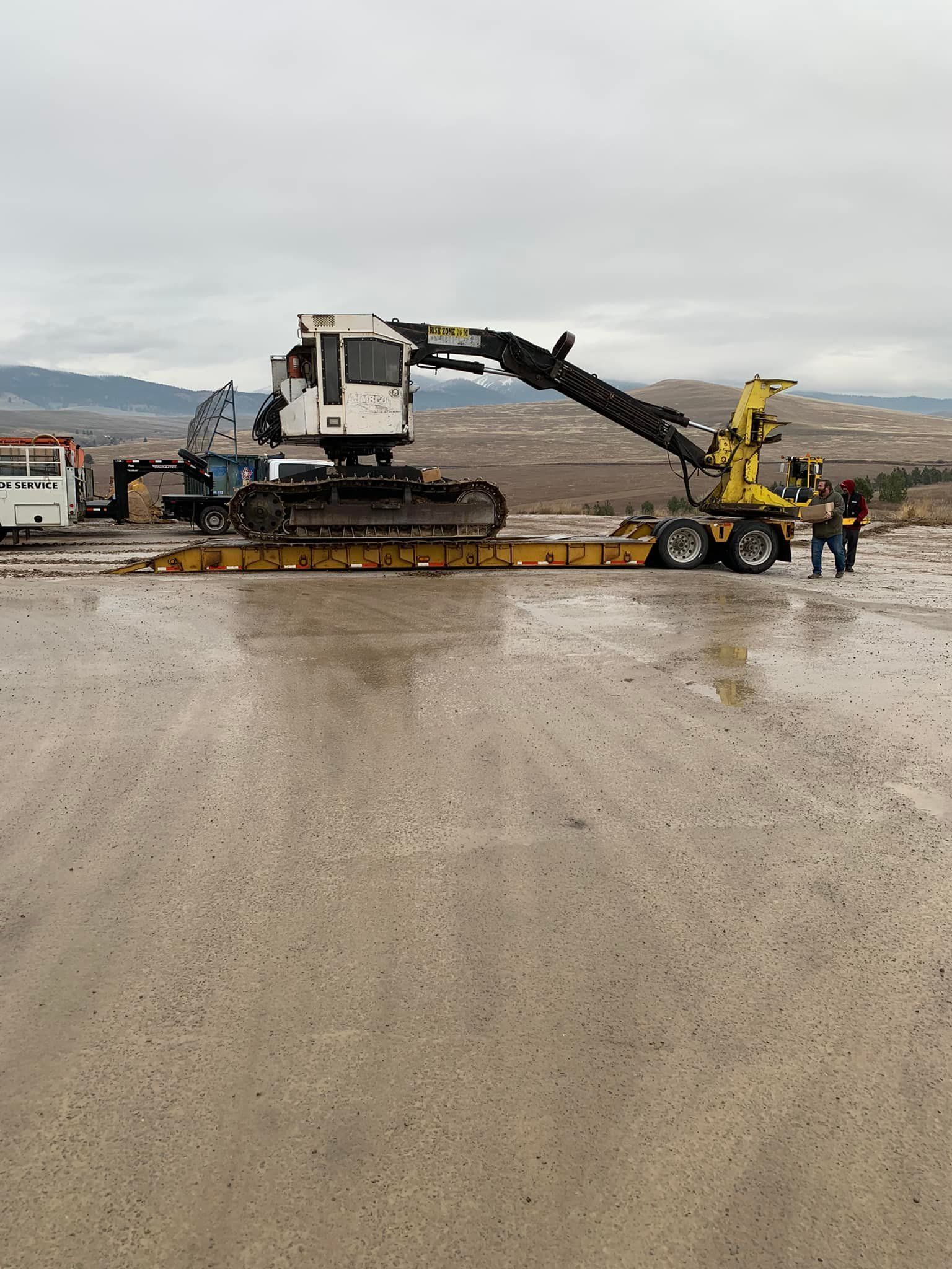Excavator on a flatbed trailer in a muddy, outdoor setting. Two people stand near the trailer.