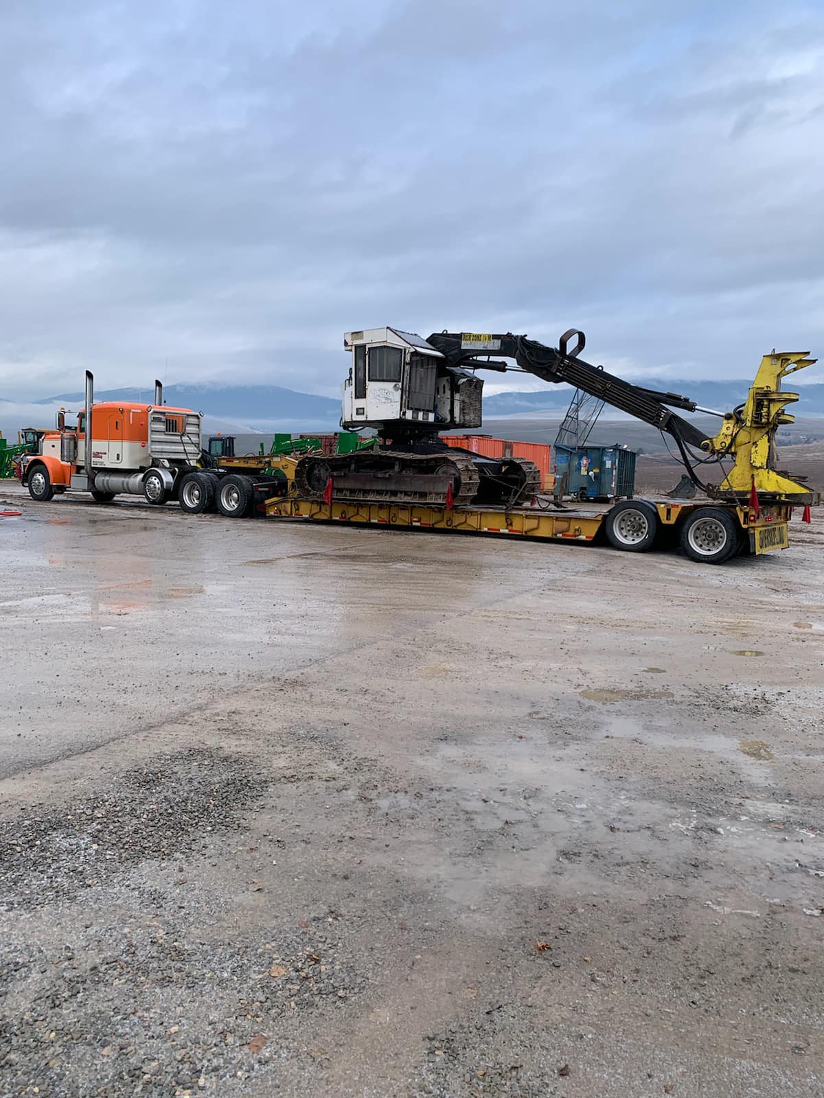 Orange semi-truck pulling a flatbed trailer carrying a large yellow forestry machine. Overcast day.