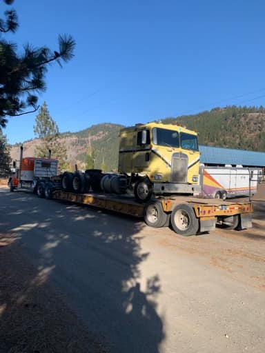 Yellow cab of a semi-truck on a flatbed trailer on a dirt road, with a partially visible truck in the background.