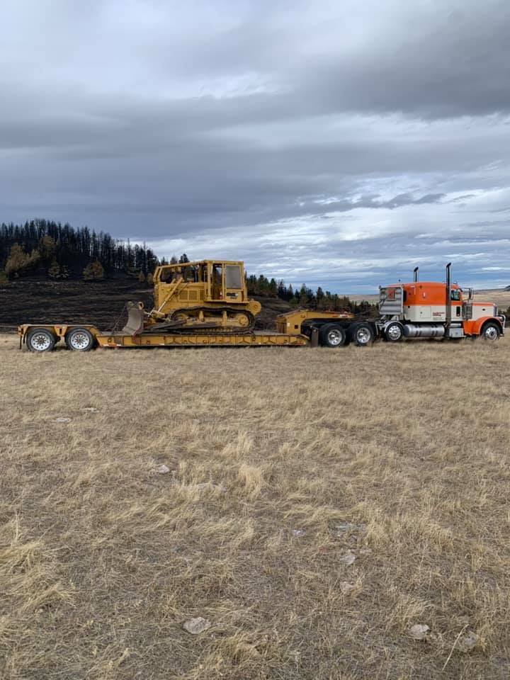 A yellow bulldozer is on a flatbed trailer pulled by an orange and white semi-truck in a field.